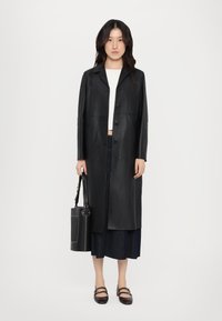 Zwarte leren lange jas met knoopsluiting, voorzien van een klassieke kraag en twee zijzakken. Model houdt een ronde handtas vast.