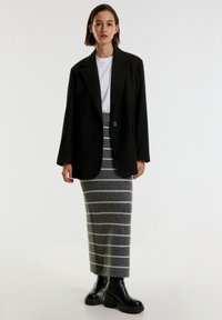 Zwarte oversized blazer over een witte t-shirt, gecombineerd met een grijs gestreepte gebreide rok. Zwarte chunky hakken ronden de outfit af.
