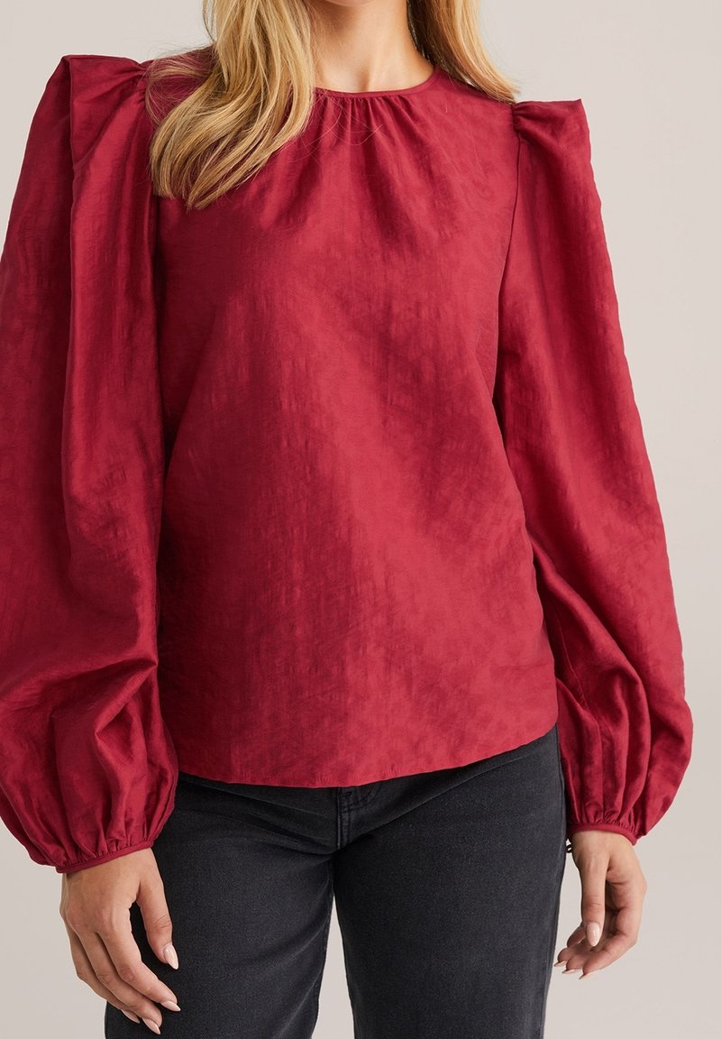 Femme portant un chemisier rouge à manches longues bouffantes avec des poignets élastiques, associé à un pantalon noir sur un fond neutre.
