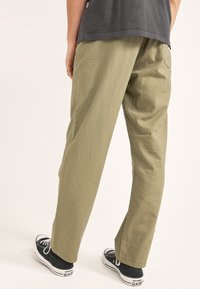 Pantalones de pierna ancha color caqui hechos de una tela texturizada, con un detalle de bolsillo trasero, combinados con zapatillas negras de corte bajo.