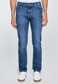 Férfi kék, karcsúsított farmernadrágban és fehér tornacipőben, sima, világos háttér előtt áll.