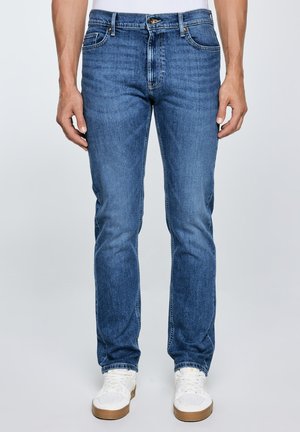 Hombre con jeans ajustados azules y zapatillas blancas de pie frente a un fondo claro y sencillo.