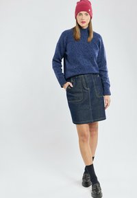 Pull marin, jupe en denim avec poches avant, chaussettes noires et chaussures épaisses, sur fond clair. Bonnet en tricot rouge comme accent.