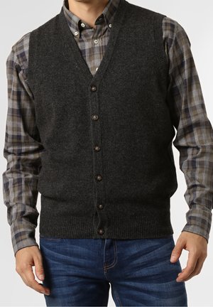 Homme portant un gilet en laine gris foncé boutonné par-dessus une chemise à carreaux marron et gris, avec un jean bleu, debout devant un fond uni.