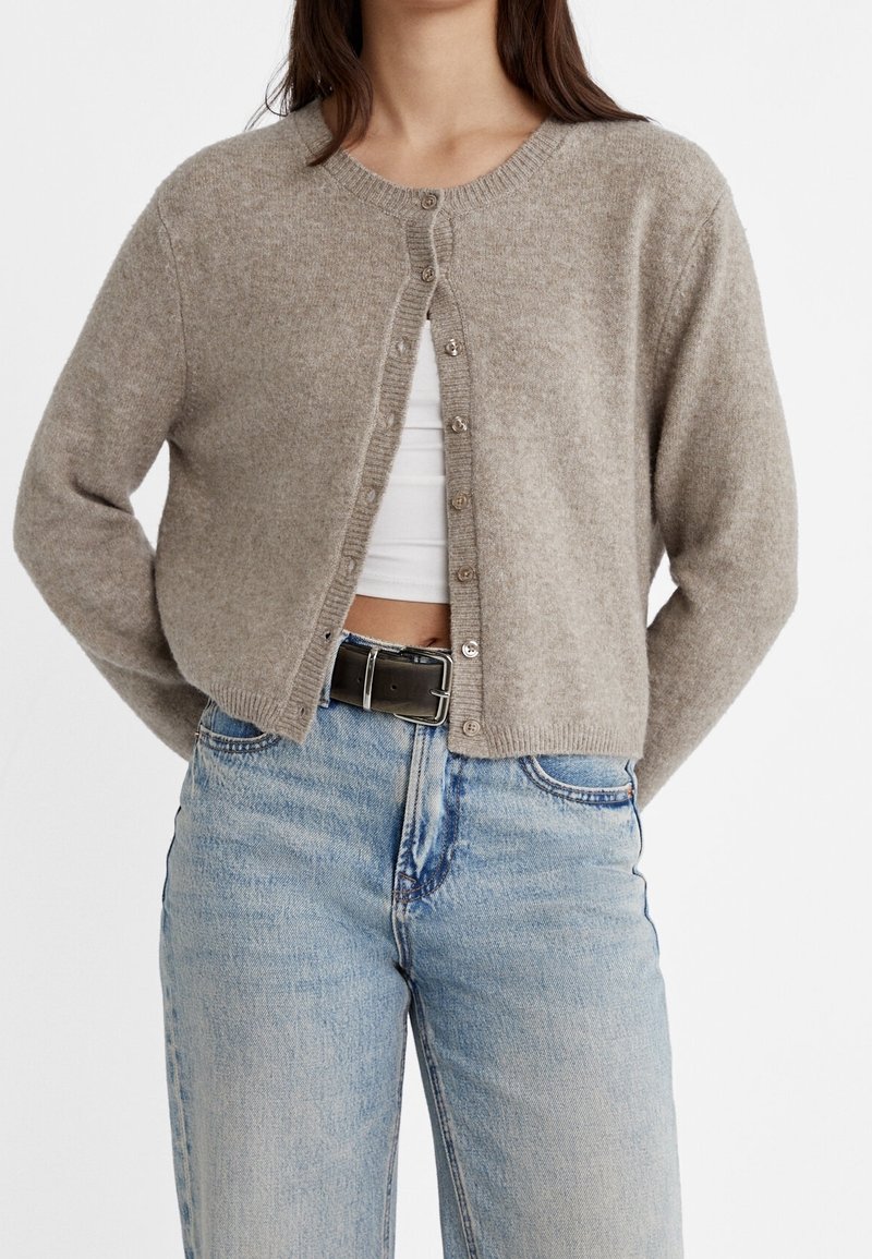 Femme portant un cardigan beige boutonné sur un crop top blanc, associé à un jean bleu clair et une ceinture noire.