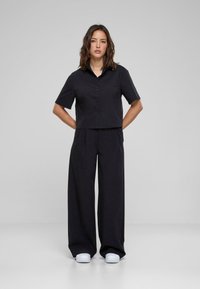 Maglietta nera cropped con maniche corte e dettagli di bottoni abbinata a pantaloni neri larghi. Il modello indossa scarpe bianche, in piedi contro uno sfondo grigio.