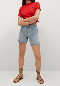 Czerwony skrócony t-shirt z logo, zestawiony z jasnoniebieskimi dżinsowymi szortami. Całość dopełniają brązowe sandały na paski.
