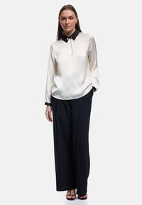 Blusa de crema satinado con un collar negro que cuenta con un acento de perla, combinada con pantalones anchos negros y sandalias negras con tiras.