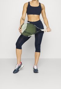 Marineblauw sportieve crop top en capri leggings, met een tennisracket met een groen ruitpatroon, en marineblauwe en rode sportschoenen.