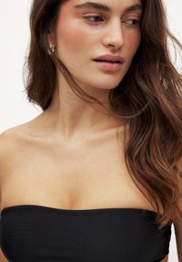 Čierny bandeau top s hladkou, elastickou textúrou. Modelka má dlhé, vlnité hnedé vlasy a nosí jednoduchý kovový náušník.