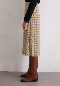 Jupe midi beige avec un motif géométrique bleu, faite d'un tissu léger ; associée à des bottes en cuir marron jusqu'aux genoux avec des détails de boucles.