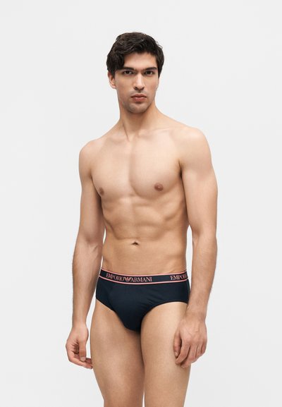 Κομψός νέος άνδρας με σκούρα μαλλιά, που φοράει μαύρα εσώρουχα Emporio Armani με ροζ λωρίδα μέσης με το λογότυπο, στέκεται μπροστά σε απλό φωτεινό φόντο.