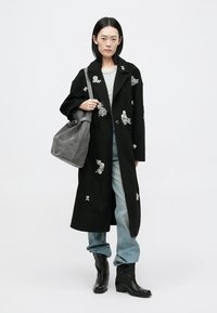 Μαύρο μακρύ παλτό με λουλουδάτες διακοσμήσεις, συνδυασμένο με γκρι τσάντα, ανοιχτό γκρι μπλουζάκι και φαρδιά μπλε τζιν, φορεμένο με μαύρα μποτάκια αστραγάλου.