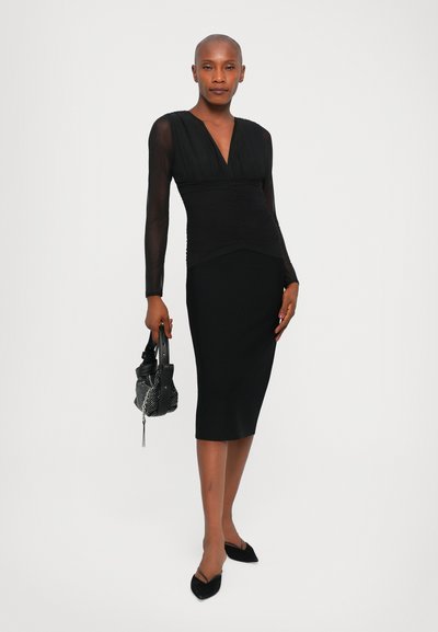 Μαύρο φόρεμα με διαφανείς μακρυές μανίκες, εφαρμοστό μπούστο και μίνιμαλ φούστα μέχρι το γόνατο. Η μοντέλο κρατάει μια μικρή μαύρη τσάντα χειρός με μεταλλικές λεπτομέρειες.