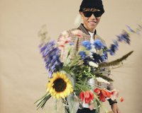 Person holding a bouquet with sunflowers, tulips, and blue flowers. They wear a checkered bomber jacket, black beanie, and sunglasses.