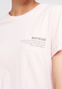 Světle růžové tričko s krátkým rukávem, na kterém je nápis "RECYCLED" a popis látky vyrobené z recyklovaných bavlněných zbytků na hrudi.