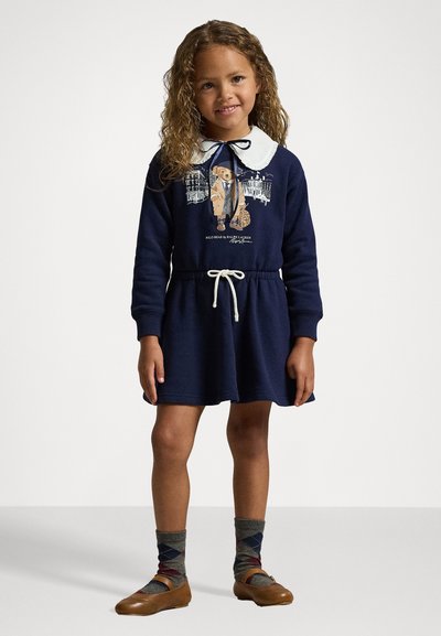 Mornarsko modra obleka z dolgimi rokavi, belim ovratnikom in vrvico za regulacijo pasu. Ima grafični vzorec medveda v obleki. Uporabljena z vzorčastimi nogavicami.