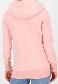 Donna che indossa una felpa leggera rosa con polsini e orlo a coste, abbinata a jeans blu, mostrata di spalle su uno sfondo neutro.