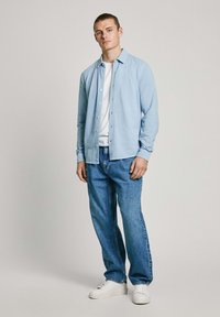 Camisa azul clara con botones, llevada sobre una camiseta blanca, combinada con pantalones de mezclilla azules y zapatillas blancas. Diseño simple y casual.