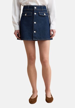 Frau trägt einen dunkelblauen, geknöpften Mini-Denimrock mit zwei vorderen Klappentaschen, eine weiße Bluse und braune flache Schuhe.