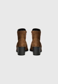 Bottines en cuir suédé marron avec un talon noir solide et des détails de zip à l'arrière, présentées sur un fond gris uni.