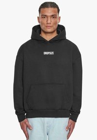 Schwarzer Kapuzenpullover aus weichem Material, mit einer Vordertasche, gerippten Bündchen und einem weißen Logo in der Mitte der Brust.