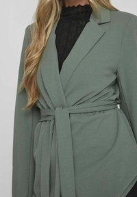 Grön kavaj i slätt tyg med bälte i midjan, strukturerade slag och långa ärmar. Svart texturerad underlag synlig.