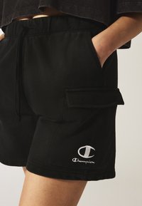 Pantaloni sportivi neri con cintura elastica, tasche laterali e una tasca cargo. Presentano un logo Champion ricamato bianco sulla gamba.