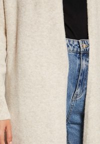 Beige, mjuk stickad cardigan med öppen framsida och ribbade ärmslut, i kombination med högmidjade blå denimjeans med knappstängning och ljus tvätt.