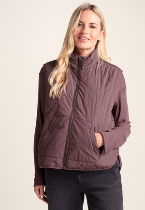 Blonde woman wearing a mauve quilted zip-up vest with pockets over a long-sleeve shirt and dark jeans, smiling with one hand in a pocket.