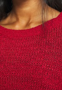 Nahaufnahme eines Halses und Schlüsselbeins einer Person, die einen roten, gestrickten Pullover mit lockerer Strickstruktur und sichtbaren dunklen Haarsträhnen trägt.