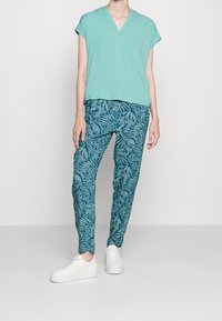Tyrkysové tričko s výstřihem do V a krátkými rukávy, spolu s modrými kalhotami s vzorem listů. Bílá teniska dotváří celý outfit.