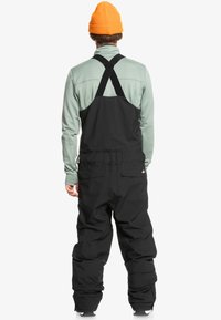 Pantalones de porteo negros con tirantes ajustables en forma de cruz, combinados con una camiseta de manga larga verde claro y un gorro de punto naranja.