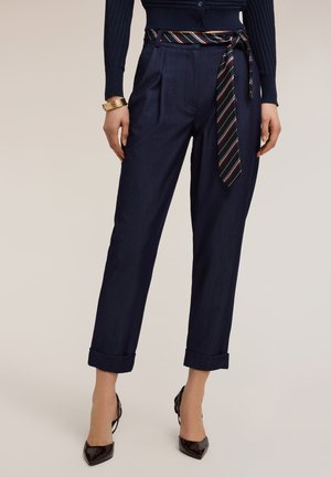 Donna che indossa pantaloni cropped blu navy a vita alta con risvolti arrotolati, cintura in tessuto a righe, tacchi neri a punta e braccialetti dorati al polso sinistro.