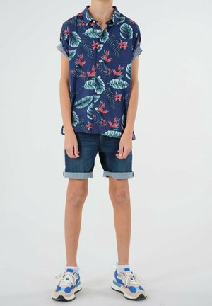 Granatowa koszula z krótkim rękawem z kwiatowymi i liściastymi wzorami, zestawiona z dżinsowymi szortami. Obuwie z niebieskimi i kremowymi akcentami, do tego białe skarpetki.