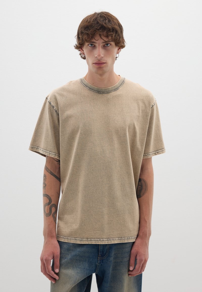 Maglietta a maniche corte di colore tan, realizzata in morbido cotone, con scollatura rotonda e dettagli di cucitura a contrasto. Indossata con jeans in denim blu.