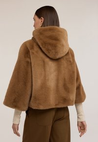 Brązowa sztuczna kurtka futrzana z kapturem, krótki krój, miękka tekstura i szerokie rękawy. Modelka nosi pod spodem jasnobeżowy sweter.