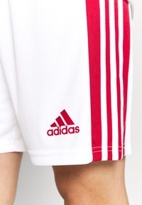 Weiße Sportshorts mit roten seitlichen Streifen und drei weißen Streifen. Mit rotem Adidas-Logo nahe dem Saum. Glatte, leichte Stofftextur.