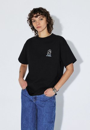 Młoda osoba z kręconymi włosami, ubrana w czarną koszulkę z napisem „Dreams of Utopia” i niebieskie dżinsy, z rękami w kieszeniach, stoi na jednolitym tle.