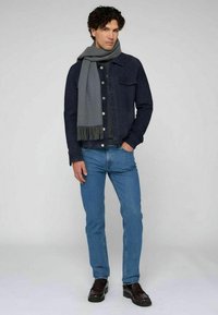 Chaqueta de mezclilla azul oscuro, bufanda gris con flecos, camisa negra, vaqueros azul claro y mocasines marrones. Atuendo casual con texturas en capas.