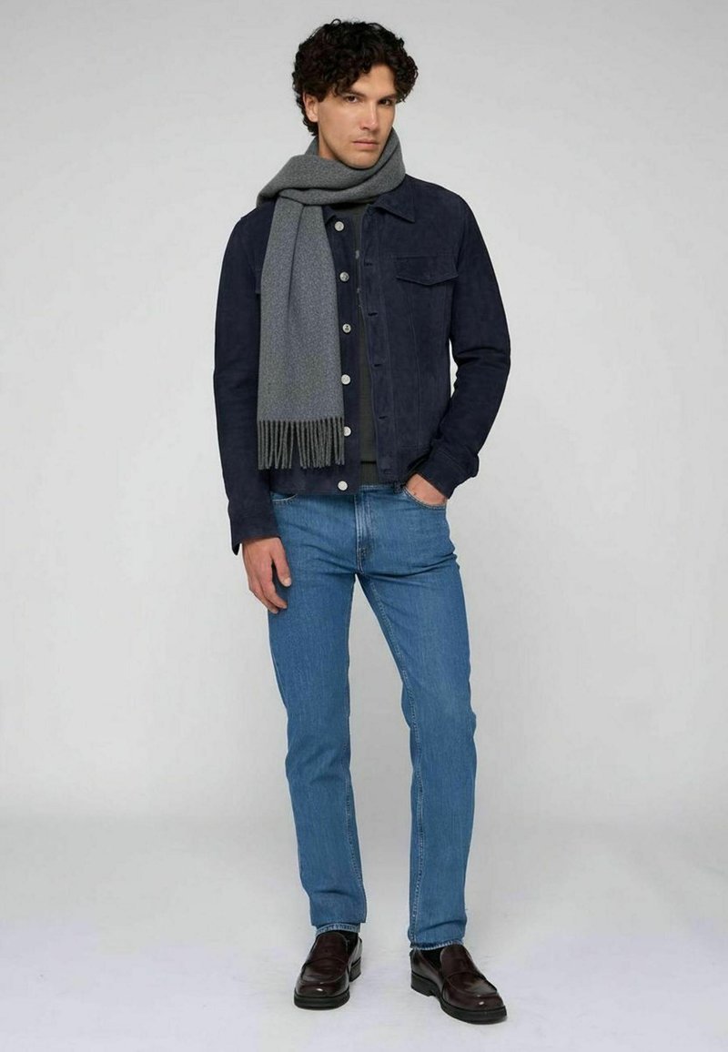 Chaqueta de mezclilla azul oscuro, bufanda gris con flecos, camisa negra, vaqueros azul claro y mocasines marrones. Atuendo casual con texturas en capas.