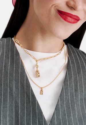 Goldene Halsketten übereinander getragen, mit einem strukturierten Anhänger an einer Kette und einem kleinen Charm an der anderen. Getragen über einem grauen Nadelstreifen-Top.