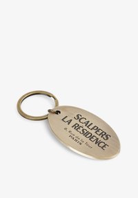 Llavero de latón en forma ovalada con texto grabado: "SCALPERS LA RÉSIDENCE 8, Rue de la Tour PARÍS." Incluye un anillo de metal para su sujeción.
