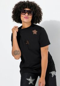 Sort T-shirt med tre stjerneformede, gennemsigtige mesh-applikationer; parret med mørke denim-materialer med istykkede stjernelapper. Model bærer solbriller.