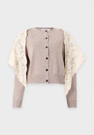 Un mannequin pose avec un pull taupe boutonné, une écharpe en dentelle crème nouée sur les épaules, un jean à jambes larges et des talons noirs.