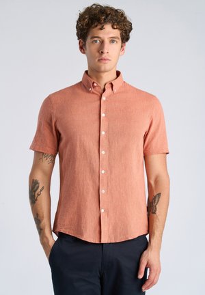 Młody mężczyzna z kręconymi włosami ubrany w terakotową koszulę na krótki rękaw z zapięciem na guziki oraz ciemne spodnie, stoi na tle jasnego, jednolitego tła.