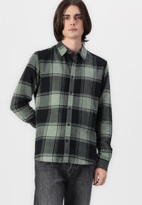 Camisa de franela de cuadros verde y negra con cuello botón, mangas largas y cuadros contrastantes. Textura suave y corte estándar.