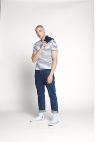 Stribet kortærmet skjorte, blå denim jeans, hvide høj-top sneakers. Rullede jeansmanchetter, navy stof draperet over skulderen, neutral baggrund.