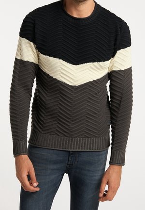 Mann in einem gerippten Strickpullover mit Zickzackmuster in Schwarz, Creme und Dunkelgrau, kombiniert mit blauen Jeans, steht vor einem einfarbigen Hintergrund.
