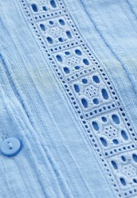 Tissu bleu clair avec des rayures verticales et un panneau central de broderie en dentelle carrée près d'un seul bouton assorti.